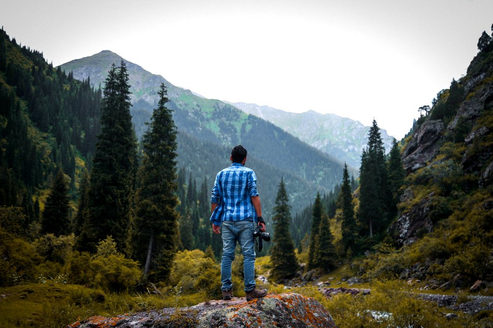 Man In Blue And White Checked Quarter-sleeved Shirt Holding Black Camera Standing On Rock