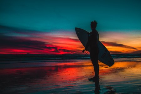 Silhouette Of Man In Wet Suit Holding White Surfboard While Standing On Beach During Golden Hour photo