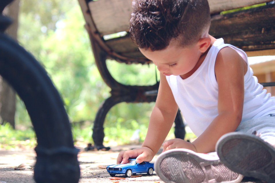 Boy In White Tank Top Playing Blue Coupe Die Cast Near Brown Wooden Bench Chair During Daytime
