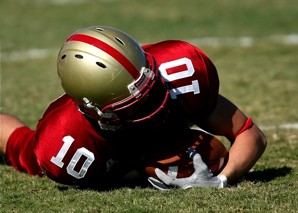 Man In Red White Football Jersey And Grey Red Helmet Holding Football And Lying On Green Grass Field