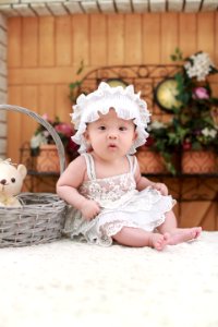Baby Wearing White Headband And White Lace Floral Dress Sitting Beside Gray Wicker Basket photo