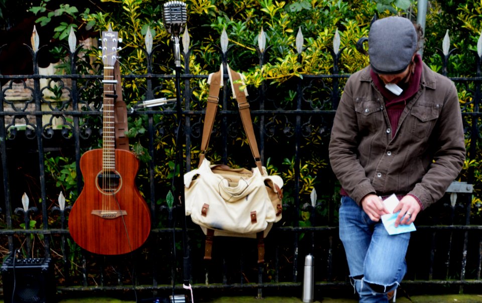 Man Leaning Against Black Steel Fence Beside White And Brown Sling Bag And Brown Acoustic Guitar