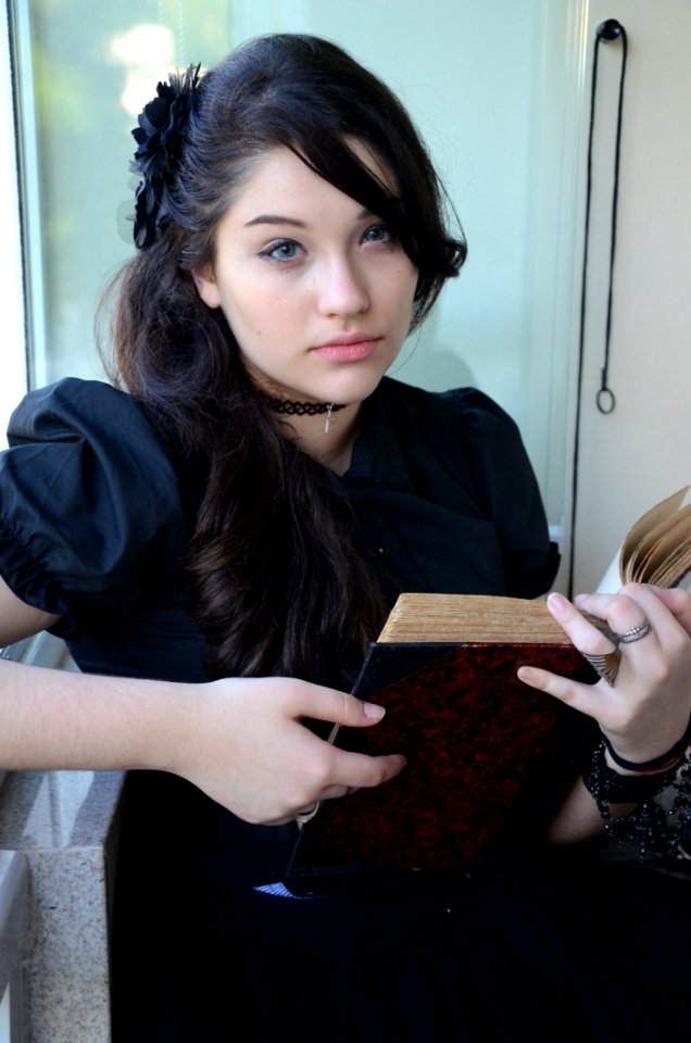 Woman With Black Hair And Wearing Black Cap Sleeve Dress Holding Book With Two Hands