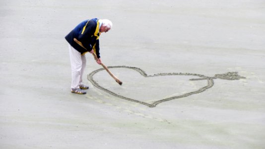 Man Wearing Blue Jacket Holding A Brown Stick Towards The Heart Drawn On Sand