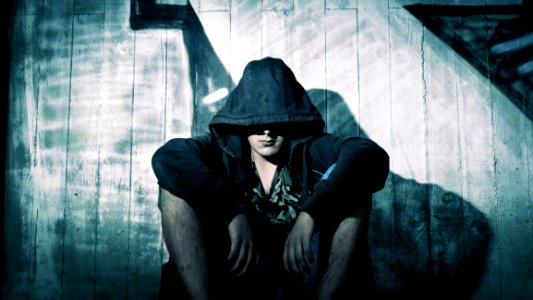 Person Wearing Black Zip Hoodie Sitting In Front Of Gray Wooden Plank Wall During Nighttime photo