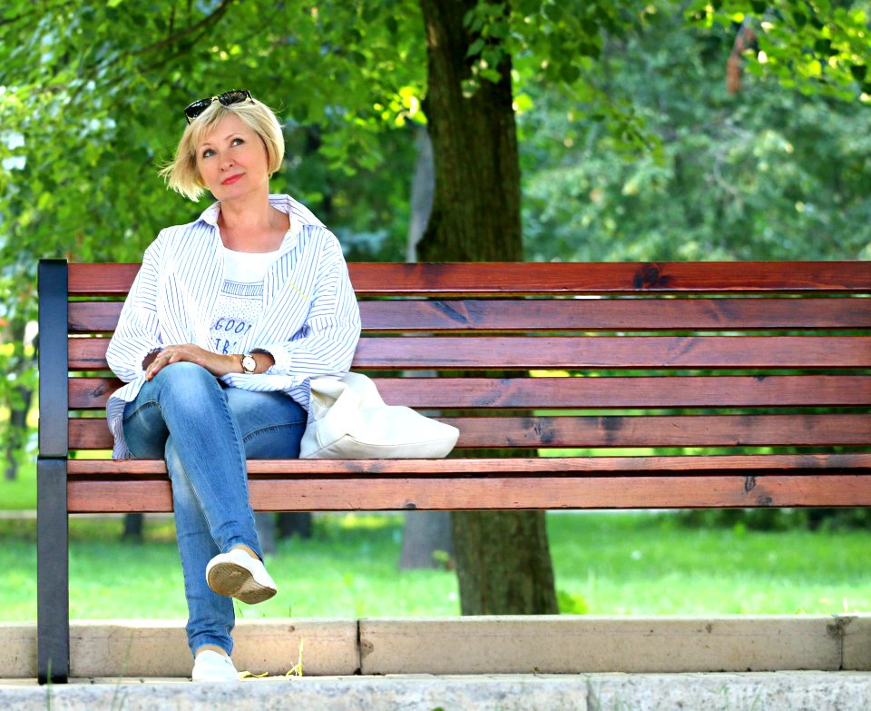 Thinking Woman In White Jacket And White Scoop Neck Shirt Blue Denim Jeans Sitting On Brown Wooden Bench Beside Green Trees During