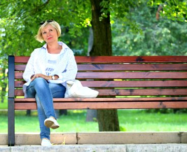 Thinking Woman In White Jacket And White Scoop Neck Shirt Blue Denim Jeans Sitting On Brown Wooden Bench Beside Green Trees During photo