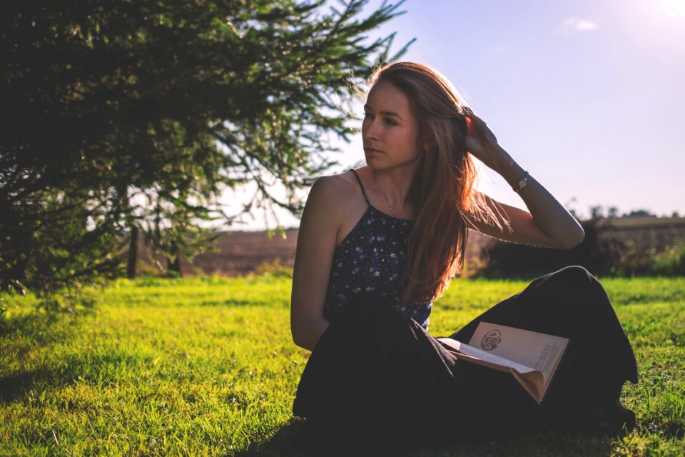 Woman In Black Tank Top And Holding Brown Book Sitting On Grass Under Clear Sunny Sky