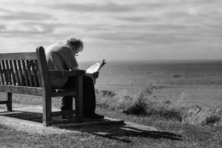 Grayscale Photo Of Man Sitting On Brown Wooden Bench Reading News Paper During Day Time photo