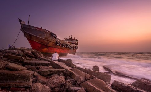Red Blue And Black Photo Of A Ship And A Man Sitting On A Stone Near Seashore photo