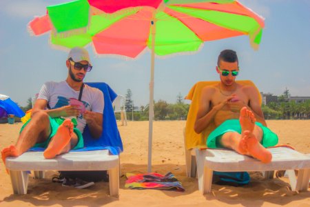 2 Man Sitting On White Beach Lounger With Green And Pink Patio Umbrella In Between