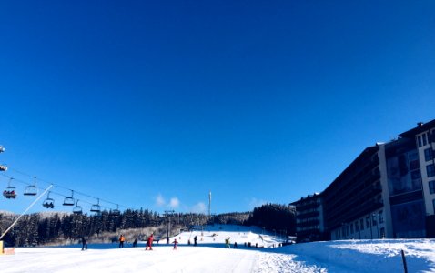 People On A Snowy Ski Hill With A Lift On The Left And A Hotel On The Right photo