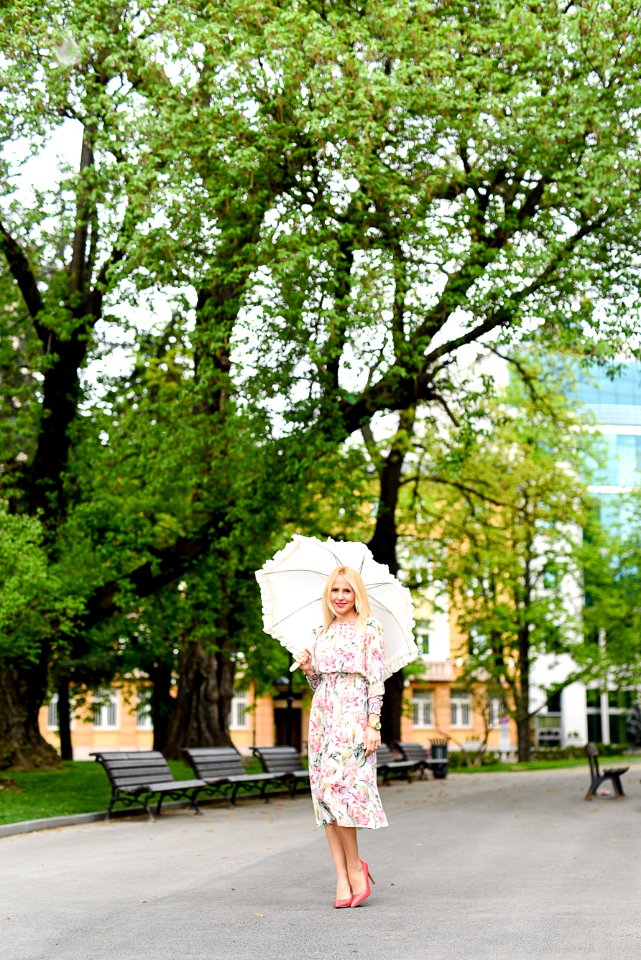 Woman In White Green And Red Floral Holding White Umbrella On Gray Concrete Pathway Near Brown Wooden Bench Near Trees During Dayt