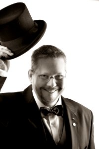 Man In Tuxedo And Eyeglasses Holding Top Hat In Grayscale
