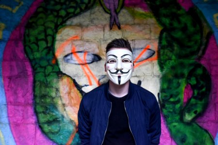 Man In White Mask In Black Crew Neck Shirt And Blue Zip Up Jacket Infront Graffiti Wall photo