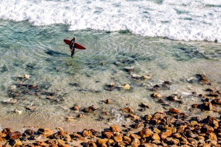Person Holding Red Surfing Board In Clear Water Near Brown Stone During Daytime