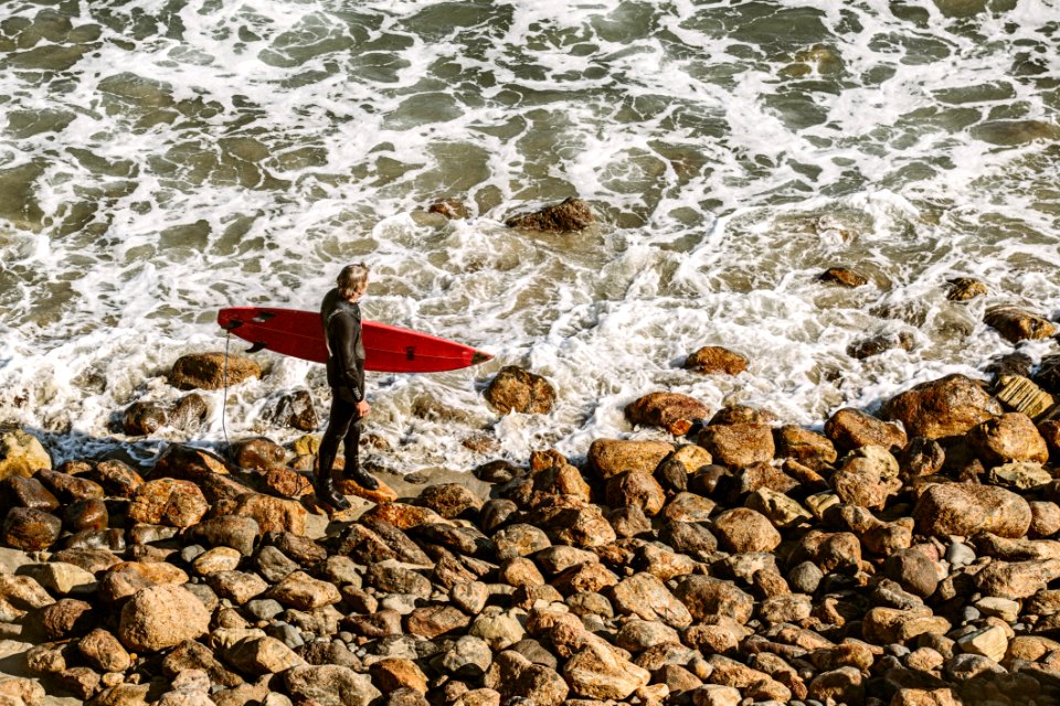 Man Wearing Wetsuit And Holding Red Surfboard On Shore