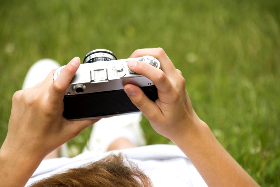 Person In White Dress Holding Grey And Black Camera On Grassland
