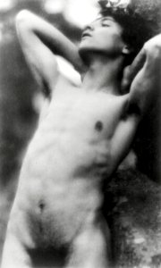 Nude photography of naked man: Portrait Photograph of nude youth with laurel wreath against rock with hands behind neck, cropped at thighs (1907) by Fred Holland.