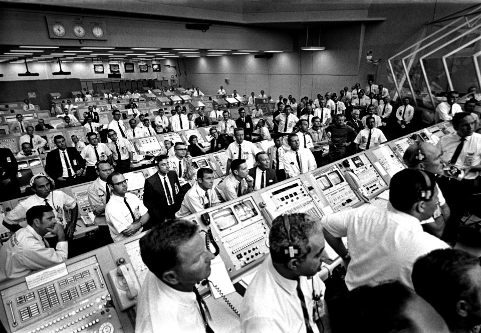 Members of the Kennedy Space Center government-industry team rise from their consoles within the Launch Control Center to watch the Apollo 11 liftoff through a window.