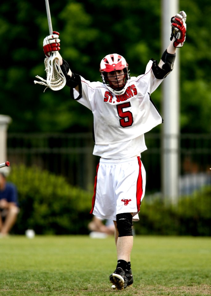 Man Holding White Lacrosse Stick Standing On Green Grass Field During Daytime