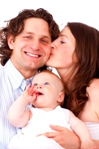 Brown Haired Woman Kissing Man In Blue White Dress Shirt Holding Baby In White Dress photo