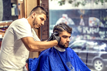 Barber In White Shirt Trimming Mans Hair In Blue Textile While Sitting Nearby Glass Window