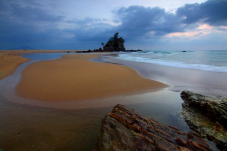 Sea Wave Crashing On Seashore With View Of Rock Formation With Trees On Top Of It Under Grey Clouds And White Sky