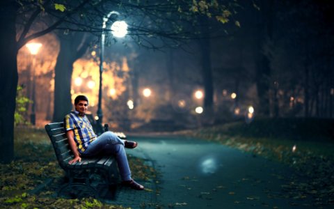 Man In Blue Denim Jeans Sitting Down By Wooden Bench Near Post Lamp Lighted