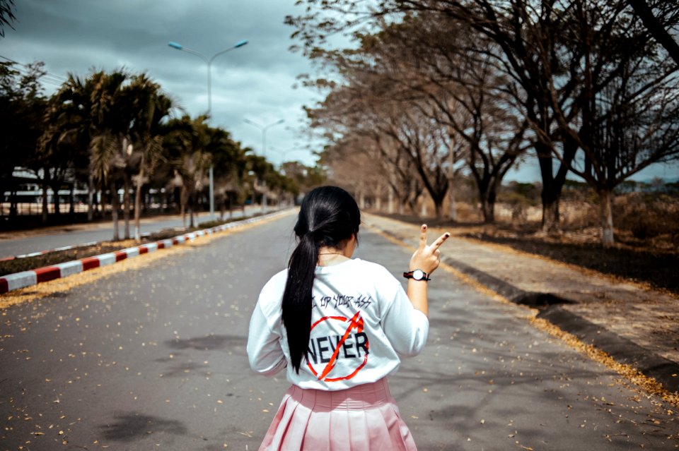 Woman Wearing White Long-sleeved Shirt And Pink Pleated Skirt Standing On Asphalt Road Between Green Trees Under Gray Sky At Dayti