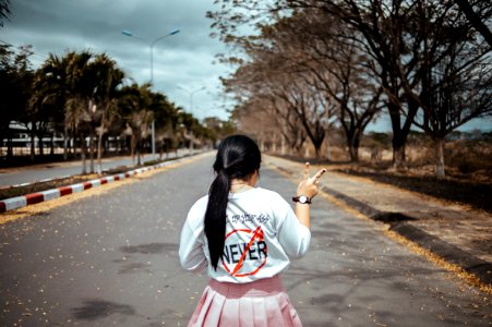 Woman Wearing White Long-sleeved Shirt And Pink Pleated Skirt Standing On Asphalt Road Between Green Trees Under Gray Sky At Dayti