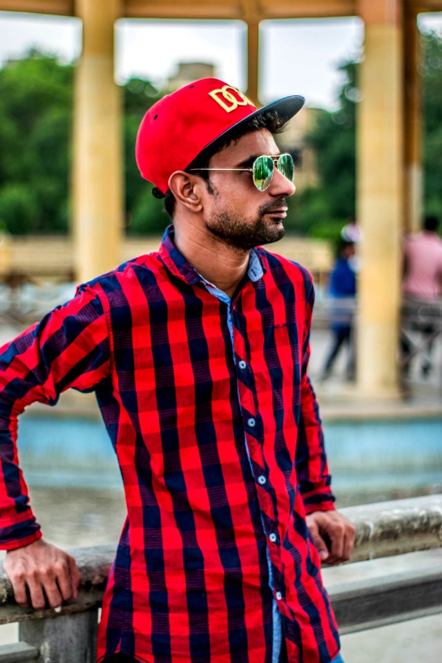 Selective Focus Photography Of Man In Red And Black Gingham Sports Shirt Leaning On Grey Concrete Fence