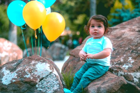 Toddler Wearing White Shirt Sitting On Rock Beside Yellow And Blue Balloons photo