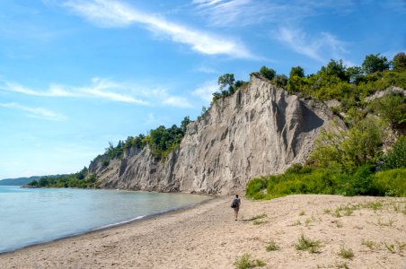 Woman Walks On Brown Seashore Near Cliff With Green Trees Under Blue And White Sky photo