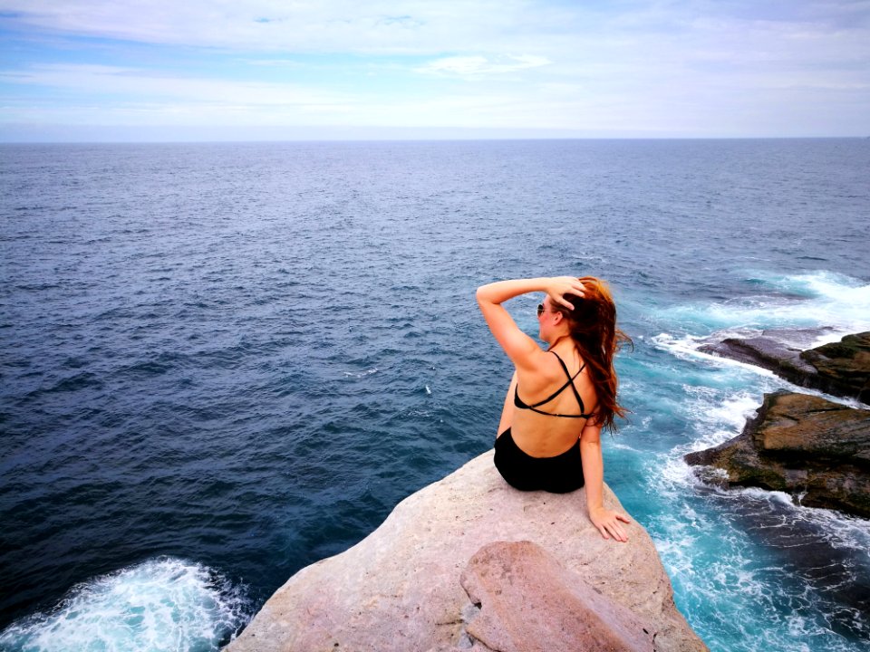 Woman In Black Swimwear Set Sitting On Top Of Gray Rocky Cliff Facing Body Of Water