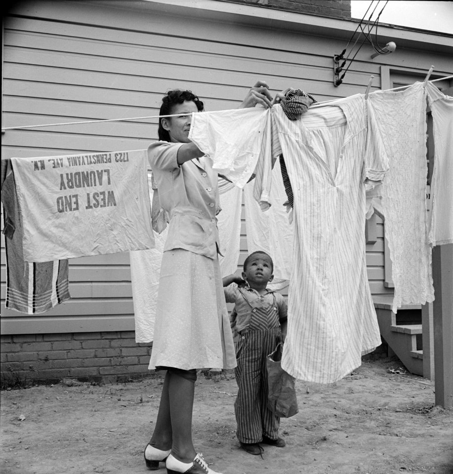Little Helper: A woman hanging out washing with her young son in front of the community building in a FSA (Farm Security Administration) housing relief camp for African Americans. Arlington, Virginia, 1942.