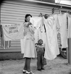 Little Helper: A woman hanging out washing with her young son in front of the community building in a FSA (Farm Security Administration) housing relief camp for African Americans. Arlington, Virginia, 1942.
