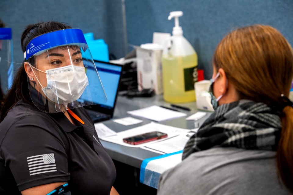 A woman wearing a mask and face shield during the COVID-19 pandemic talks with a masked woman who will be receiving a vaccine at an administration site.