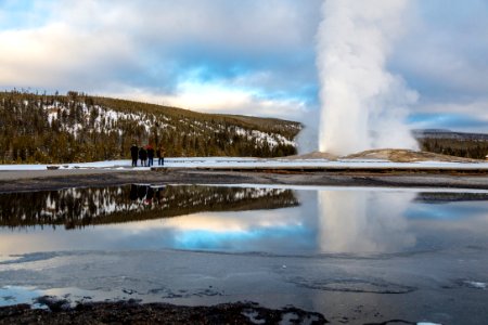 Old Faithful eruption with onlookers reflected in a puddle