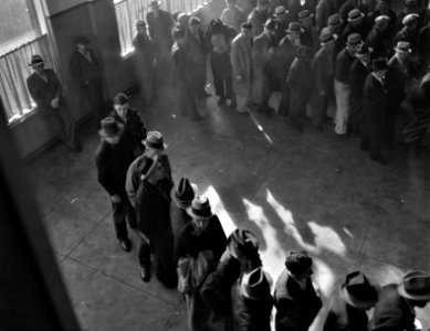 Shuffle With Tired Feet: Line of men inside a division office of the State Employment Service office at San Francisco, California, waiting to register for benefits on one of the first days the office was open. January 1938.