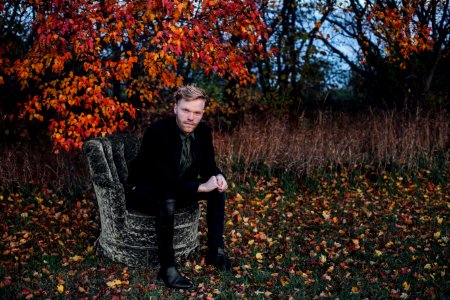 man wearing black suit sits on black concrete chair beside brown tree