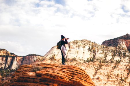 man wearing black jacket holding DSLR camera while taking photo standing on brown cliff during day time photo