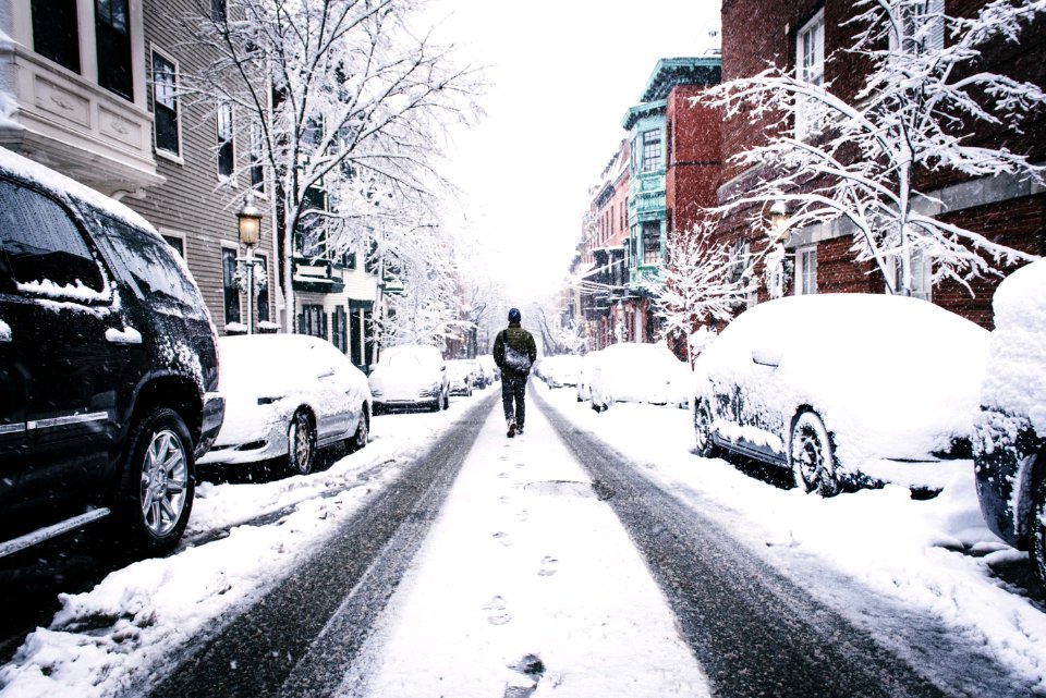 man walking on road covered with snow between vehicles and concrete buildings during daytime