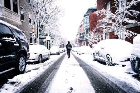man walking on road covered with snow between vehicles and concrete buildings during daytime photo