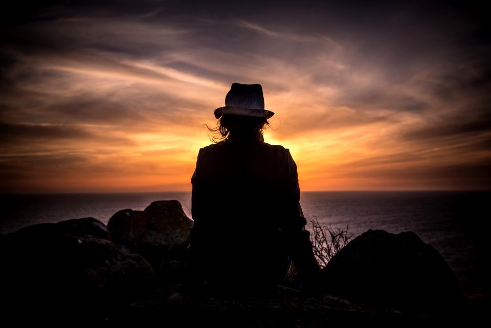 silhouette of man sitting on stone looking at body of water