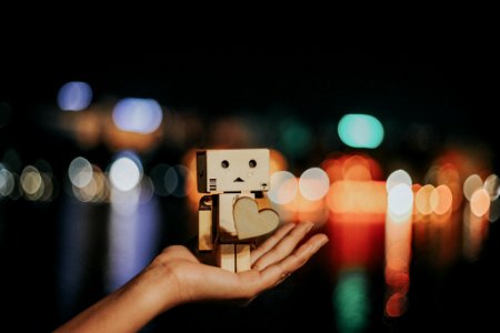 A person's hand holding up a box character with a heart, in front of a blurred out city background. photo