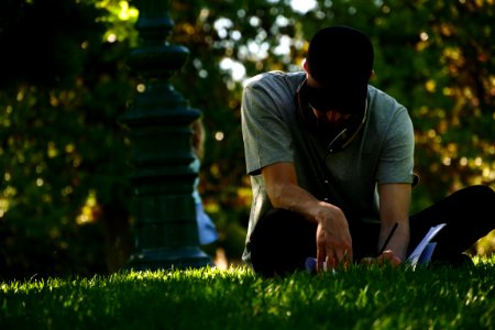 selective focus photography of man sitting on grass and writing on notebook during daytime