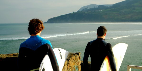 two male wearing wetsuits holding surfboards standing nearby each other on top of hill with view of beach below photo