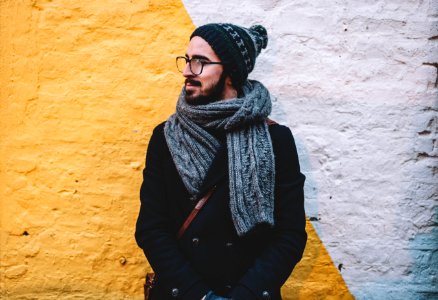 man in black coat with grey scarf standing on front of white and yellow painted wall photo