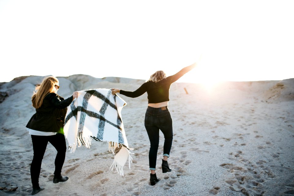 vignette photography of two woman holding scarf walking on sand